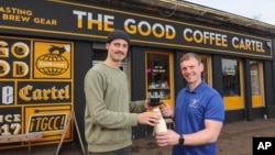 This undated handout photo shows farmer and owner of Mossgiel Organic Farm Bryce Cunningham, right, giving a bottle of milk to barista Jacob Smith, as they pose outside The Good Coffee Cartel in Glasgow, Scotland. (Mossgiel Organic Dairy via AP)