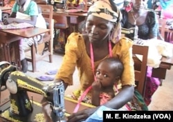 A seamstress trainee works on a sewing machine with a child on her lap.