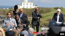 Un homme manifeste avec une balle de golf rouge marquée d’une croix gammée pendant que le candidat républicain à la présidence américaine Donald Trump, à l’arrière droite, coiffé d’une casquette blanche, est sur le point de donner un discours sur la cours de golf Trump Turnberry, en Écosse, en Grande-Bretagne, 24 juin 2016.