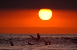 FILE - Surfers wait for waves as the sun goes down the day before the beach was scheduled to close during the coronavirus outbreak, April 30, 2020, in Newport Beach, Calif.