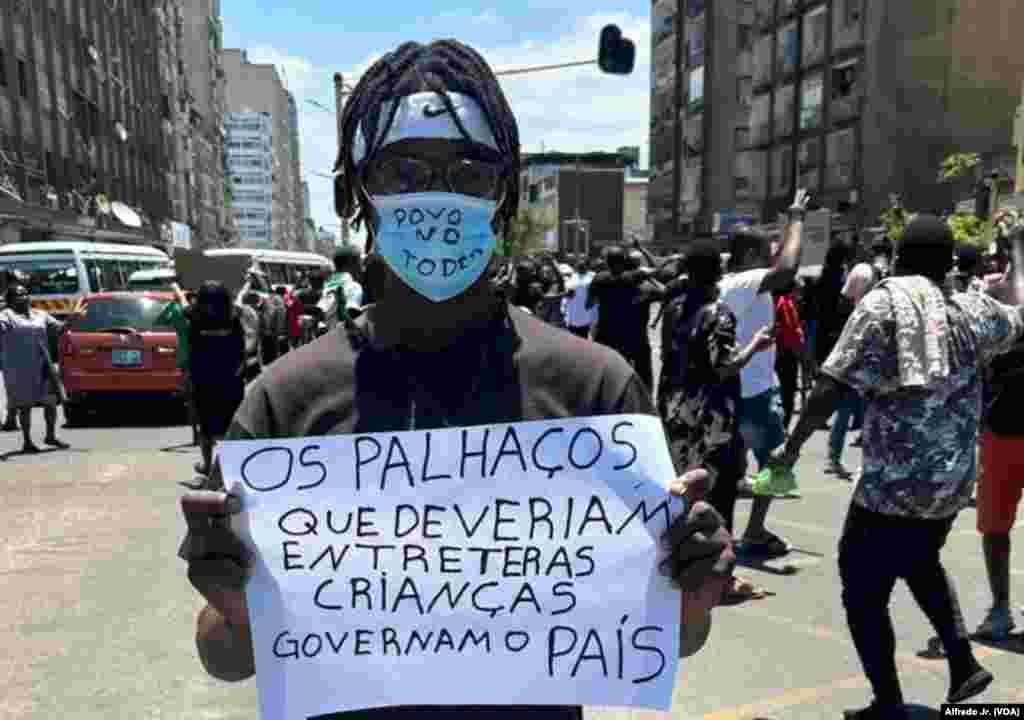 Um jovem com uma máscara a dizer “Povo no Poder” segura um cartaz a dizer “Os palhaços que deviam entreter as crianças governam o país”. Ele participa num protesto na cidade de Maputo