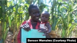 A happy mother and child display their loyalty vaccination card against the background of their farm which shows healthy crops.