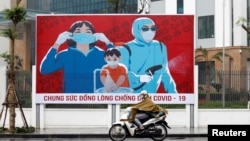 Một người phụ nữ đeo khẩu trang lái xe máy qua tấp áp phích tuyên truyền việc phòng chống virus corona (COVID-19) ở Hà Nội hôm 3/4/2020. 