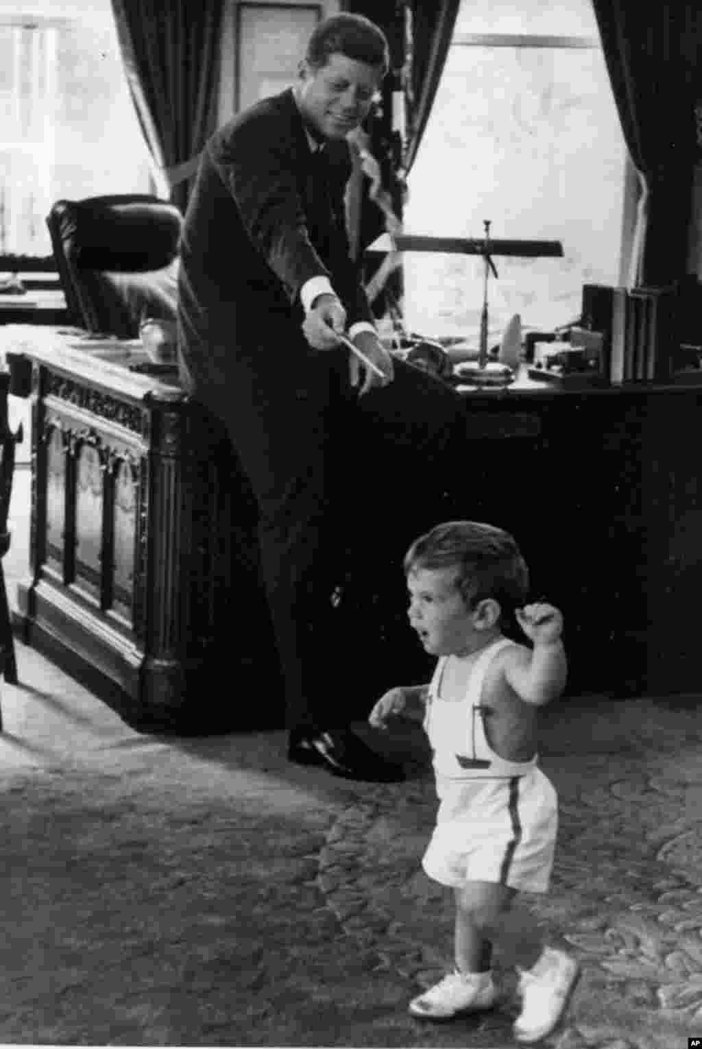 U.S. President John F. Kennedy holds out a pencil toward his 18-month-old son John Jr., who takes a few steps in the Oval Office of the White House in Washington, D.C., on Friday, May 25, 1962. (AP Photo/White House, Robert Knudsen)