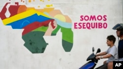 Dos venezolanos se trasladan en una motocicleta frente a una mural donde se aprecia uno de los eslóganes oficiales de defensa del Esequibo como parte del territorio de Venezuela, en el barrio 23 de Enero de Caracas, en diciembre de 2023.