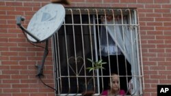 Una mujer mira desde una ventana situada junto a una antena de DirectTV, instalada en una vivienda pública en Caracas, Venezuela.