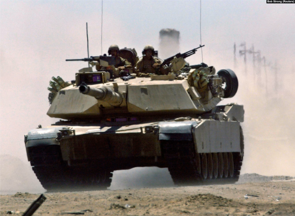 Un tanque M1A1 Abrams del Cuerpo de Marines de EEUU acelera por una carretera iraquí cerca de un puesto de control de tráfico militar en Faluya el 1 de julio de 2004. Se cree que el Ejército de EE. UU. tiene 2509 Abrams en varias versiones, con 3700 adicionales almacenados.