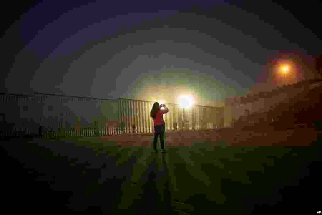 A woman records with her phone, as floodlights from the United States light up the border wall, topped with wire, along the beach in Tijuana, Mexico.