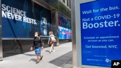 FILE - Pedestrians walk past a signs hanging outside Pfizer headquarters in New York and one hanging at a bus stop encouraging the COVID-19 booster, May 23, 2022. 