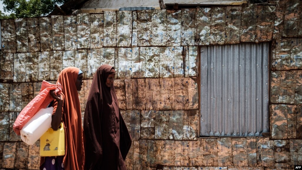 FILE - Women walk in front of a metal fence made of vegetable oil cans from USAID at the Dadaab refugee complex, northeastern Kenya, on April 18, 2018. 