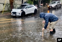 A Mission District resident attempts to unclog a few storm drains on a flooded street as a driverless Waymo taxi passes in the background, Nov. 22, 2024, in San Francisco.