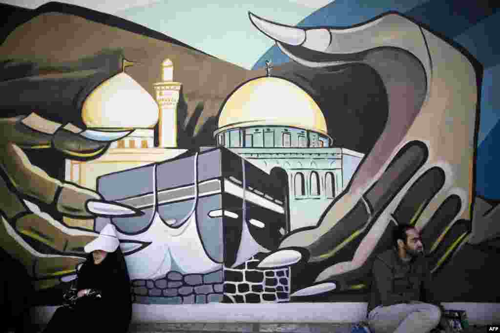 Seorang perempuan dan seorang laki-laki Iran duduk di depan sebuah mural yang melukiskan Kabah di Mekah dan Kubah Batu Yerusalem di alun-alun Palestina saat mereka menunggu datangnya Sholat Jumat dalam sebuah demonstrasi di Teheran untuk menandai Hari Quds atau Hari Yerusalem. &nbsp;
