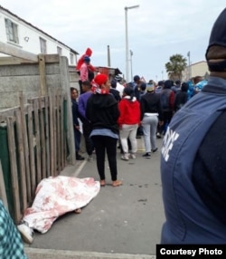 People gather near the covered body of a teenager killed in a gang shootout in Hanover Park, a township of Cape Town, South Africa, Sept. 19, 2017. A police officer stands in the foreground. (Special to VOA) Click on photo for related video.