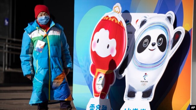 A woman wearing a face mask to protect against COVID-19 stands next to figures of the Winter Paralympic mascot Shuey Rhon Rhon left, and Winter Olympic mascot Bing Dwen Dwen on a street in Beijing, Jan. 15, 2022.