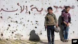 A Syrian boy holds a toy gun as he plays soccer with others between destroyed buildings with graffiti that reads "Syria al-Assad," in the old city of Homs, Syria, Friday, Feb. 26, 2016.