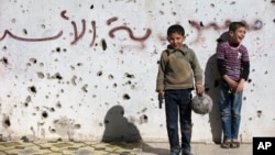 A Syrian boy holds a toy gun as he plays soccer with others between destroyed buildings with graffiti that reads "Syria al-Assad," in the old city of Homs, Syria, Friday, Feb. 26, 2016.