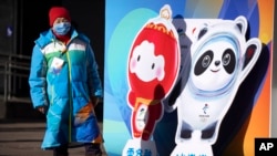 A woman wearing a face mask to protect against COVID-19 stands next to figures of the Winter Paralympic mascot Shuey Rhon Rhon left, and Winter Olympic mascot Bing Dwen Dwen on a street in Beijing, Jan. 15, 2022.