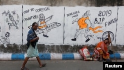 FILE - A man walks past a woman who sits by a wall with graffiti that mocks Indian politicians ahead of the general election in Kolkata, India, Apr. 1, 2019.