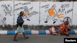 FILE - A man walks past a woman who sits by a wall with graffiti that mocks Indian politicians ahead of the general election in Kolkata, India, Apr. 1, 2019.