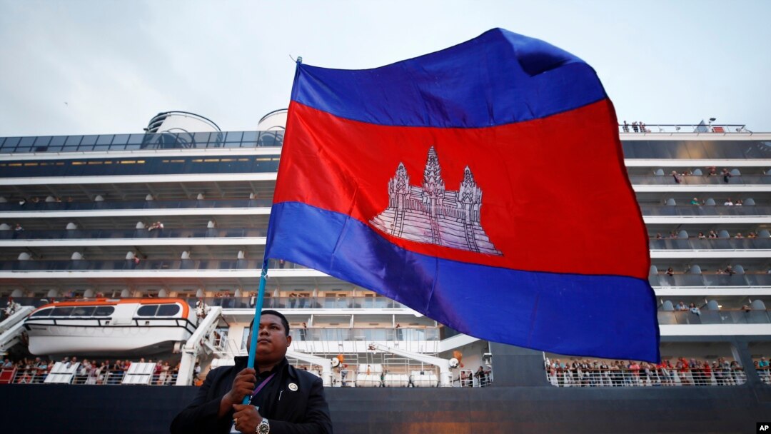 Seorang pria Kamboja memegang bendera nasionalnya di depan MS Westerdam, milik Holland America Line, berlabuh di pelabuhan Sihanoukville, Kamboja, Jumat, 14 Februari 2020. (Foto: AP)