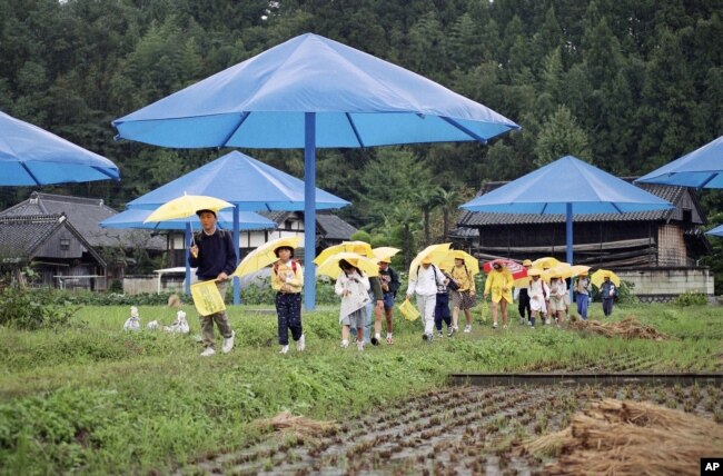 FILE - Public school students carry their own yellow umbrellas through a rice paddy dotted with giant blue umbrellas from the project by Christo in Japan's Sato River Valley, Oct. 9, 1991. (AP Photo/Mitsuhiko Sato)