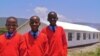 New students Tasiana, Happiness and Samwel stand in front of their new secondary school, smiling in their new school uniforms. (Red Sweater Project)