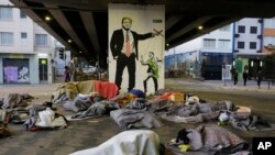 Para tunawisma tidur di bawah jembatan di depan mural yang menggambarkan Presiden A. Donald Trump sebagai dalang yang memanipulasi Presiden Brasil Jair Bolsonaro, di pusat kota Sao Paulo, Brasil, 23 Juli 2019. (Foto: AP)