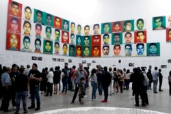FILE - People stand under the portraits of 43 college students who went missing in 2014 in an apparent massacre, by Chinese concept artist and government critic Ai Weiwei at the Contemporary Art University Museum in Mexico City, Mexico, April 13, 2019.