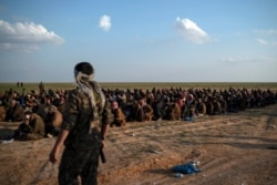 FILE - In this Feb. 22, 2019 file photo, U.S.-backed Syrian Democratic Forces (SDF) fighters stand guard next to men waiting to be screened after being evacuated out of the last territory held by IS militants, near Baghouz, eastern Syria.