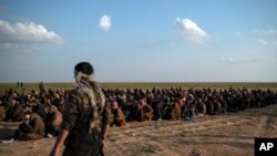 FILE - In this Feb. 22, 2019 file photo, U.S.-backed Syrian Democratic Forces (SDF) fighters stand guard next to men waiting to be screened after being evacuated out of the last territory held by Islamic State group militants, near Baghouz, eastern Syria.