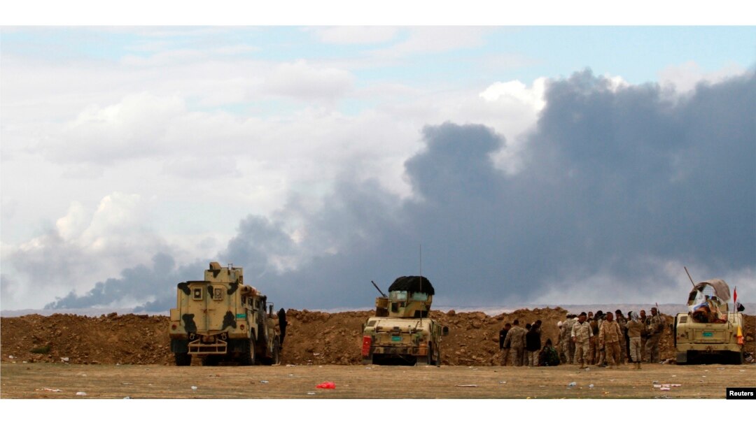FILE - Iraqi soldiers gather near vehicles as smoke rises from oil wells in the Ajil field east of Tikrit in the Salahuddin province that were set on fire by Islamic State militants in March 2015.