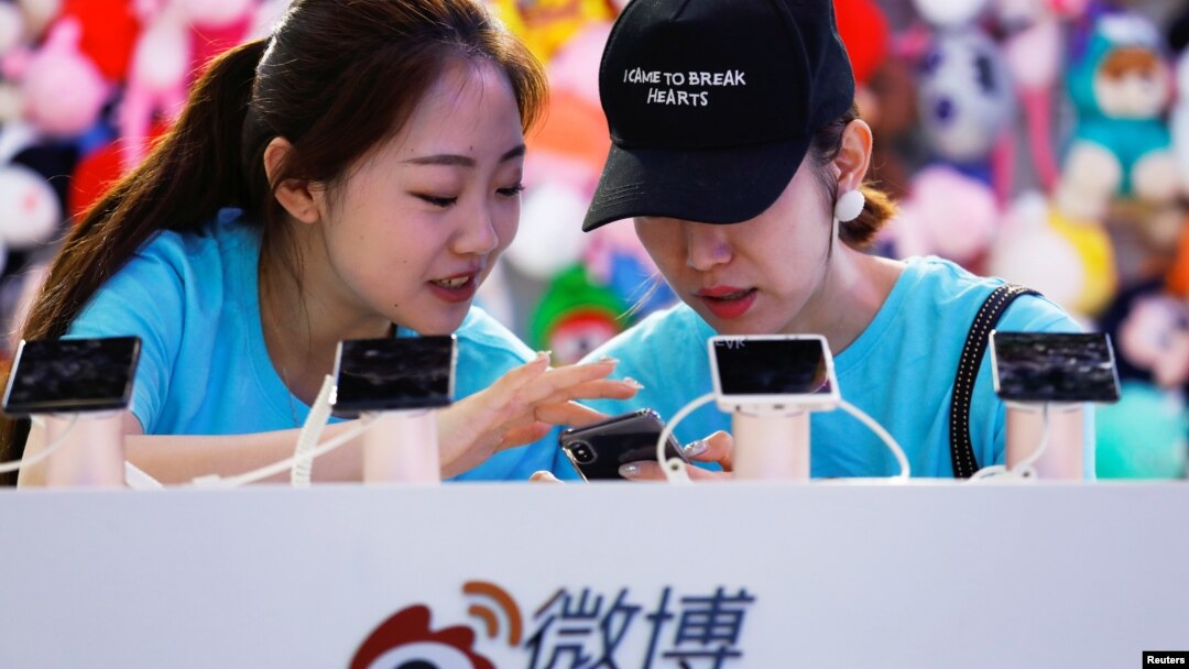  2018年4月27日，在北京舉行的全國移動互聯網大會（GMIC）期間，工作人員在新浪微博的攤位上使用智能手機。一個人的帽上有英文，意思是“我是來讓人心碎的”。 