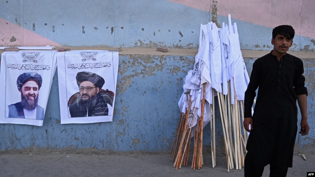 一名阿富汗商贩在街头悬挂的塔利班领导人画像旁售卖塔利班旗帜。（2021年8月27日）(photo:VOA)