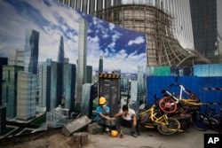 ILE- In this June 26, 2018, file photo Chinese workers take a break outside a construction site wall depicting the skyscrapers in the Chinese capital at the Central Business District in Beijing.