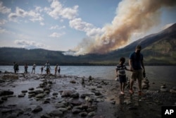 In this Aug. 12, 2018 photo provided by the National Park Service, people walk along the shore near Lake McDonald Lodge as the Howe Ridge Fire burns in Glacier National Park, Montana.