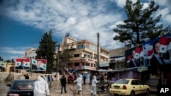 FILE - Workers spray disinfectant to prevent the spread of the coronavirus, on a street lined with billboards showing Syrian President Bashar al-Assad, in Qamishli, Syria, March 24, 2020.