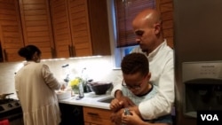Jeremiah Randall checks his father Jeremy's watch to see how long he must wait for dinner. His stepmother, Ariam Mohamed, prepares a chicken and pasta dish for iftar, a dinner during Ramadan. (C. Guensburg/VOA)