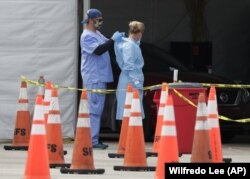 In this July 5, 2020, file photo, healthcare workers help each other with their personal protective equipment at a drive-thru coronavirus testing site outside Hard Rock Stadium in Miami Gardens, Fla.