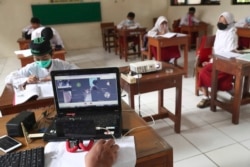 Seorang guru menggunakan laptopnya sementara siswa yang mengenakan masker duduk terpisah selama uji coba kelas dengan protokol COVID-19 di sebuah sekolah dasar di Jakarta, Jumat, 4 Juni 2021. (Foto: AP)