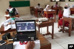 Seorang guru menggunakan laptopnya sementara siswa yang mengenakan masker duduk terpisah selama uji coba kelas dengan protokol COVID-19 di sebuah sekolah dasar di Jakarta, Jumat, 4 Juni 2021. (Foto: AP)