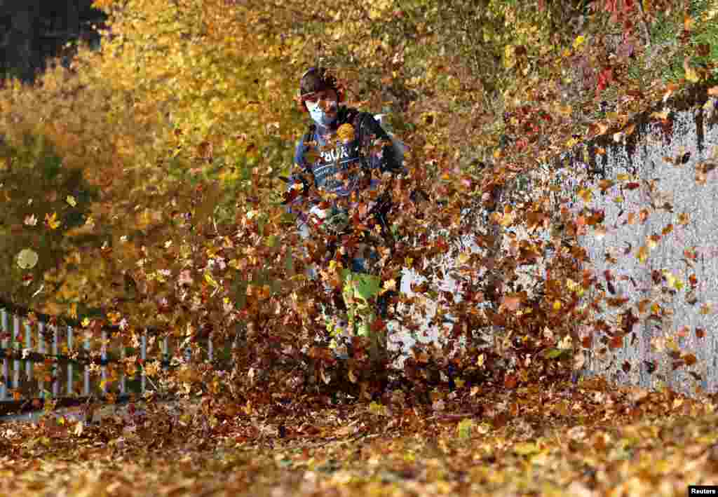 A worker wearing a protective mask uses a blower to remove leaves from a walkway during sunny autumn weather near Stallikon, Switzerland.