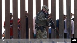 Tentara AS berjalan di sepanjang pagar perbatasan AS-Meksiko di San Diego, California, sementara para migran tampak berada di wilayah Meksiko, Minggu (25/11). 
