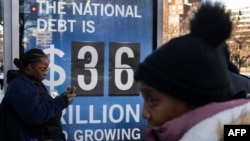 People wait for a bus near a National Debt Clock billboard display showing the United States gross national debt and each American family's share of the debt in Washington, DC, on December 30, 2024.