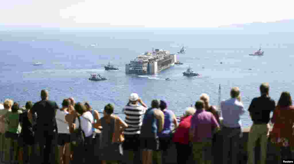 Người dân xem du thuyền Costa Concordia quay ngược chiều kim đồng hồ trong khi đang được làm nổi tại đảo Giglio, Italia, ngày 23 tháng 7, 2014. Một đoàn 14 tàu, dẫn đầu là tàu kéo Blizzard, kéo chiếc Concordia đến một cảng gần thành phố Genoa ở miền bắc Italia, nơi nó sẽ được tháo dỡ làm phế liệu.