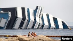 June 20, 2012: Vacationers bask in front of the wreckage of capsized cruise liner Costa Concordia near the harbour of Giglio Porto.