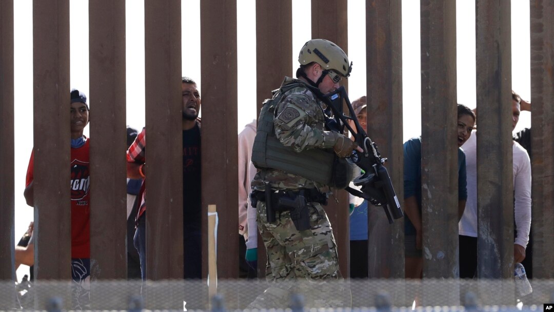 Tentara AS berjalan di sepanjang pagar perbatasan AS-Meksiko di San Diego, California, sementara para migran tampak berada di wilayah Meksiko, Minggu (25/11). 