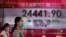 People wearing face masks walk past a bank electronic board showing the Hong Kong share index at Hong Kong Stock Exchange Monday, April 20, 2020.