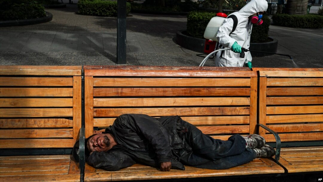 Seorang petugas kebersihan kota menyemprotkan disinfektan sementara seorang pria tidur di bangku di Plaza de Armas, di Santiago, Chili, 15 April 2020.