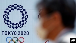 FILE - A man wearing a face mask walks passed a billboard with the logo of the Tokyo 2020 Olympics, in Tokyo, Japan, April 2, 2020.