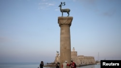 People wearing protective face masks bump elbows next to the statue of a deer at the entrance to Mandraki harbor, amid the COVID-19 pandemic, on the island of Rhodes, Greece, April 13, 2021. Island of Rhodes prepares for tourism season opening.